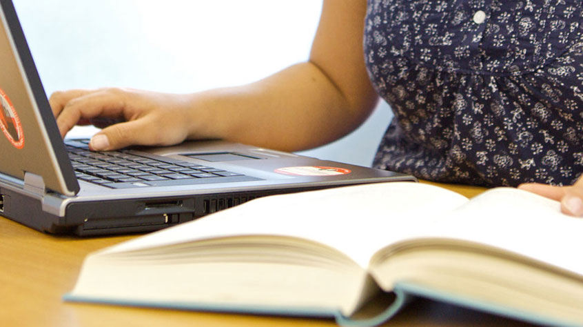Hand on laptop keyboard, with an open book on the table.