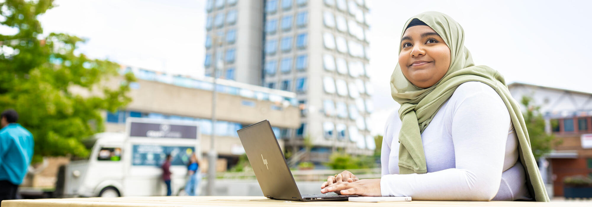A smiling woman in a light green hijab works on a laptop at an outdoor table, with leafy trees and modern campus buildings behind her.