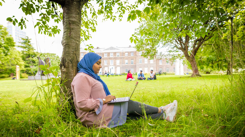 Student sitting next to a tree, working on her laptop.