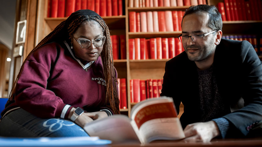 Student sitting with a lecturer in the Law library and looking at a book together.
