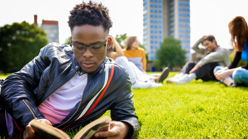 Student leaning on the ground in a park and reading a book on a sunny day.