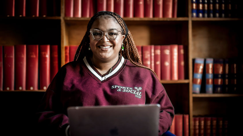 Student sitting in front of a large bookshelf, with a laptop on her knee and she is smiling at the camera.