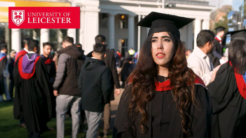 Student wearing a cap and gown, standing outside De Montfort Hall and talking to camera.
