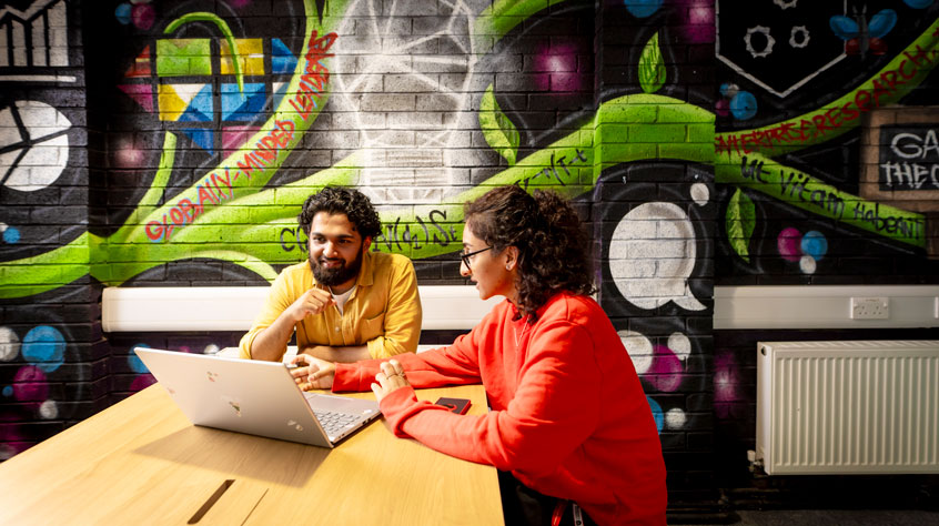 Students sitting at a table with a laptop between them and a colourful wall behind them.