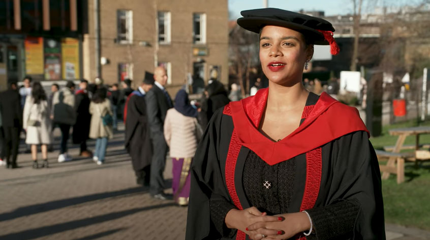 Student wearing a cap and gown and speaking to camera outside the Students' Union on Leicester campus.