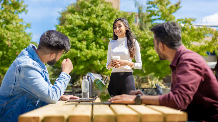 Three students talking around a wooden bench on campus