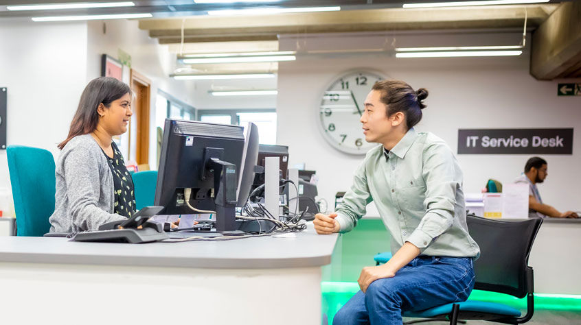 Staff member talking to a student with a desk between them in the library.