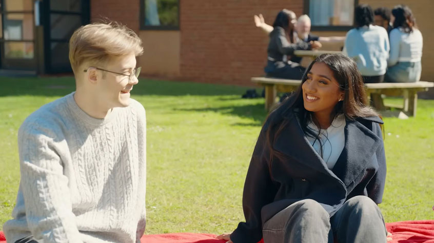 Two students sitting on the grass, on a sunny day, talking to each other.