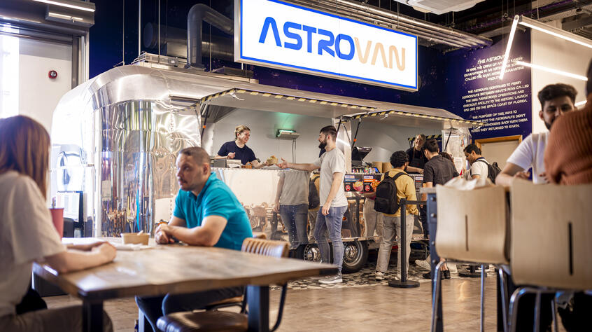 A social space with people sitting at tables talking and an Astro Van food truck serving people. 