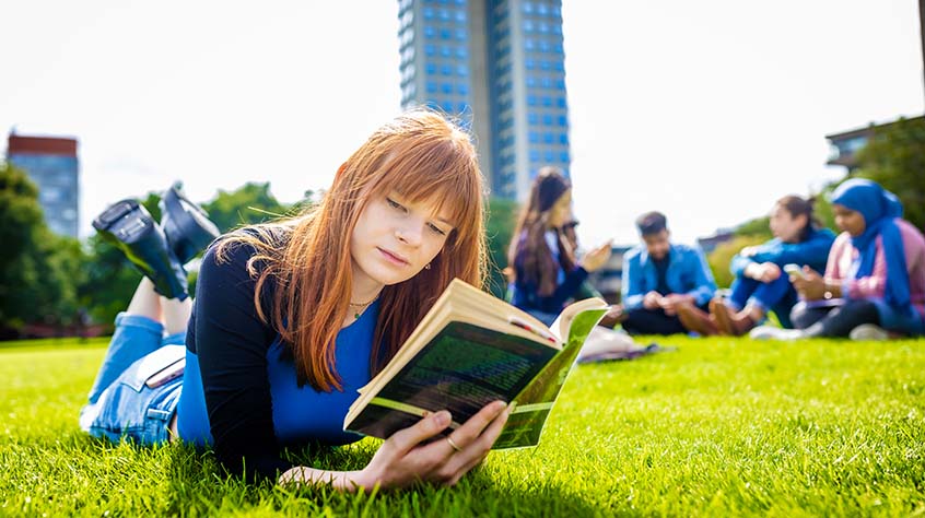 Student lying on their front reading a book in a park