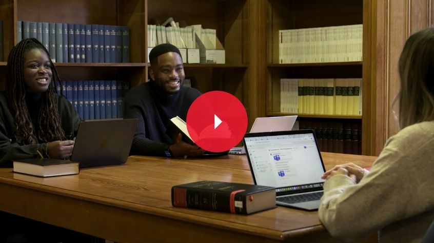 Two students sat down at a table with a third person just out of view on a laptop