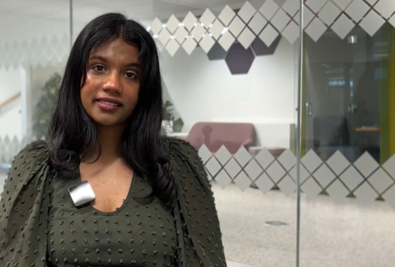 Smiling student seated to the left, facing camera directly, with a glass wall in the foreground