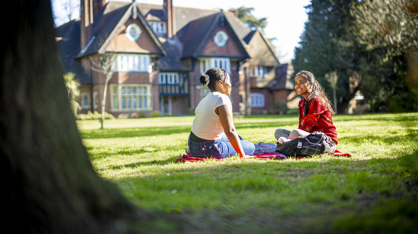 Two female students sitting on green grass sharing a picnic.