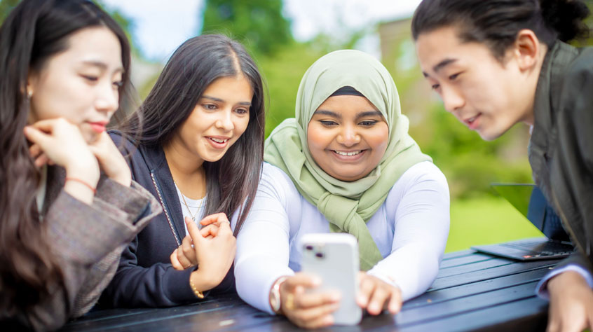 Students sitting on a bench gathered around a phone, taking a selfie