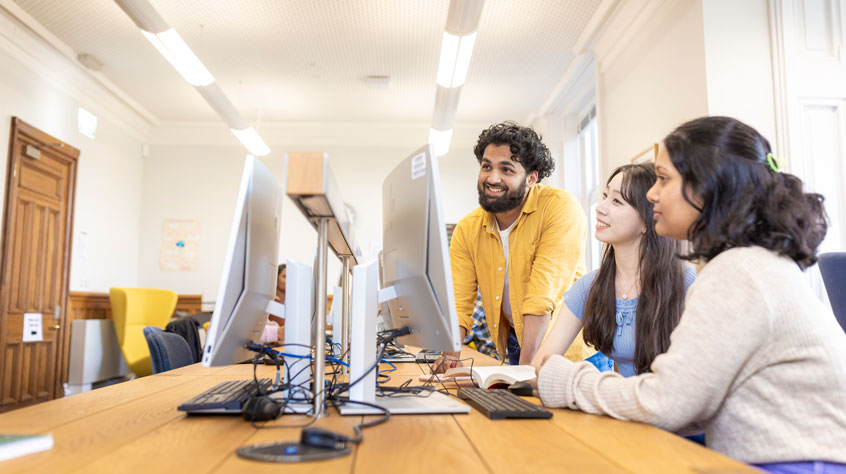 Students sitting on a table with computers, gathered around one of them.