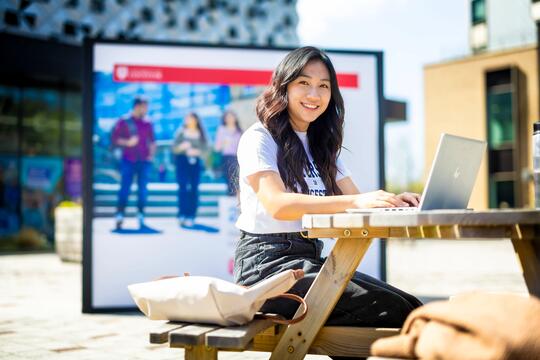 Student outside on campus bench in sunshine facing and smiling 