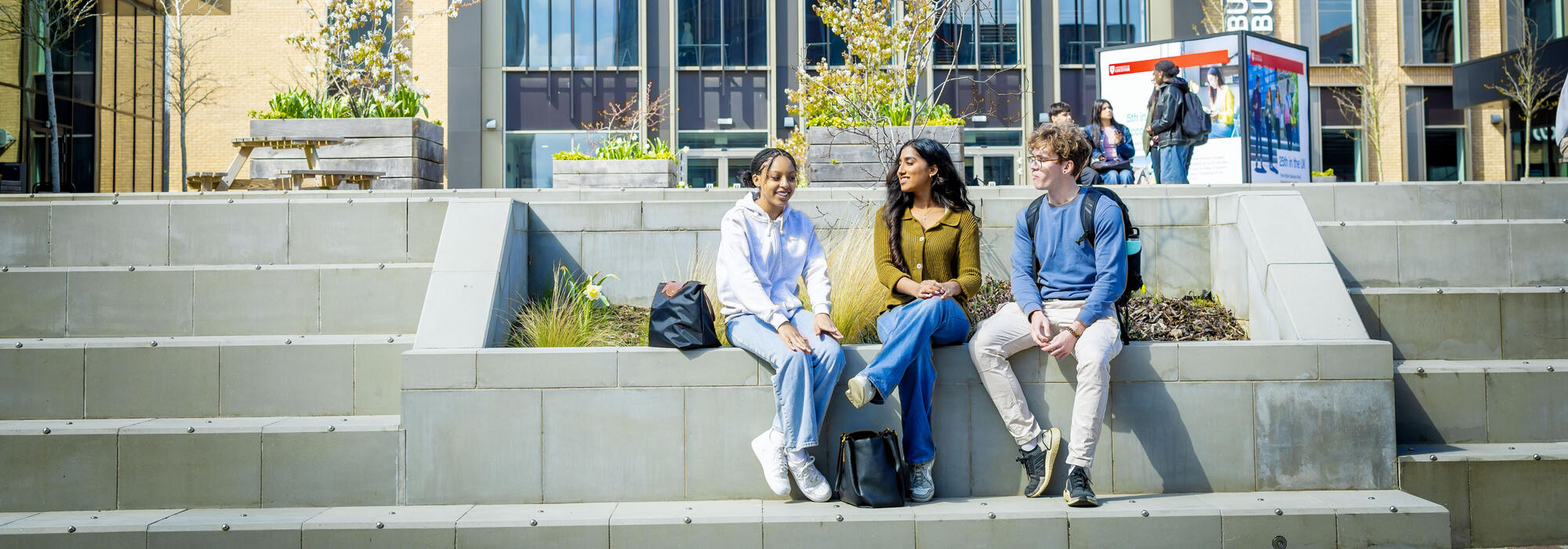 Three university students sit on concrete steps outdoors on a sunny day.