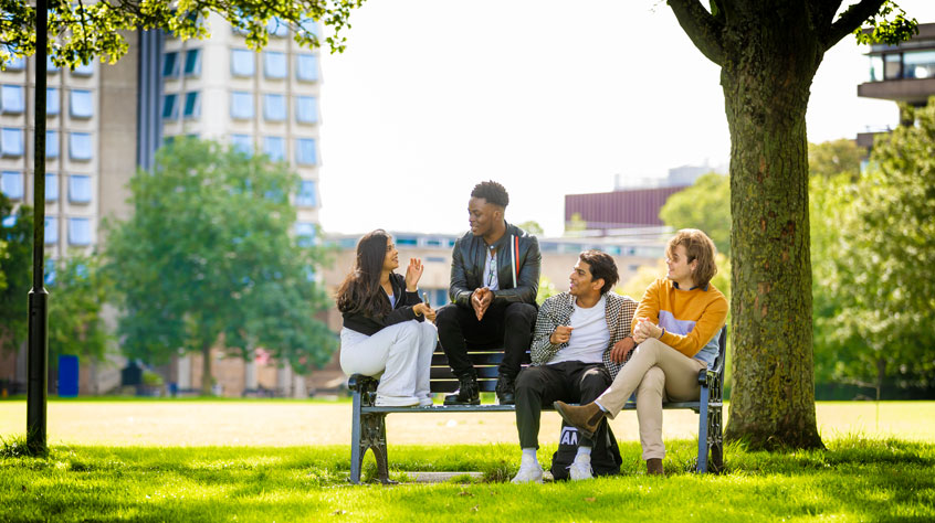 Four students sitting on a bench in Victoria park on a sunny day