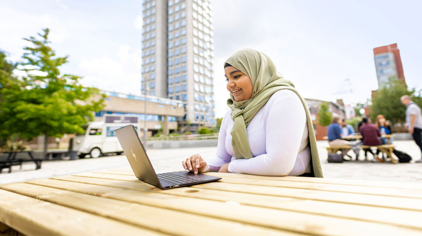 Female student sitting outside on a bench, smiling whilst browsing on a laptop.