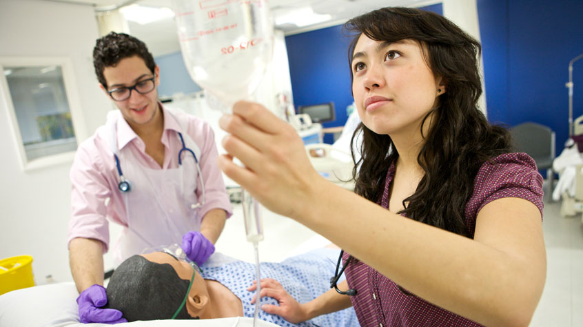 Medicine student putting up an IV drip whilst another checks the status of a medical dummy.