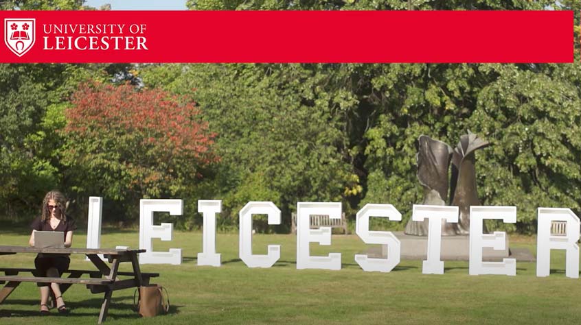 Person sitting on a bench on campus in front of a big Leicester sign.