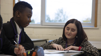 Boy sat at a desk in the classroom with a teacher helping him.