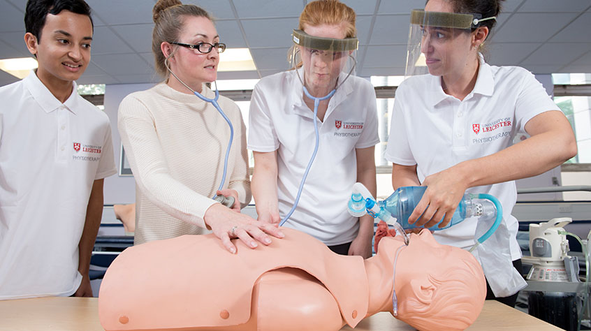 Four students in light coloured clothing, two with stethoscopes around their necks, practising a medial procedure on a dummy