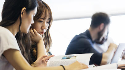 Two students engaged in studying together at a classroom desk, surrounded by books.