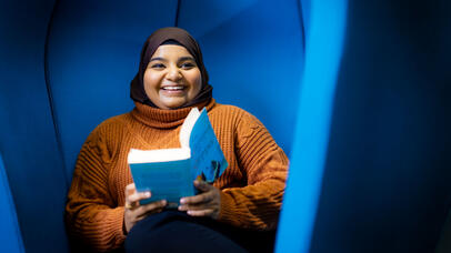 A woman in a hijab sits in a chair, smiling joyfully while holding a book in her hands.