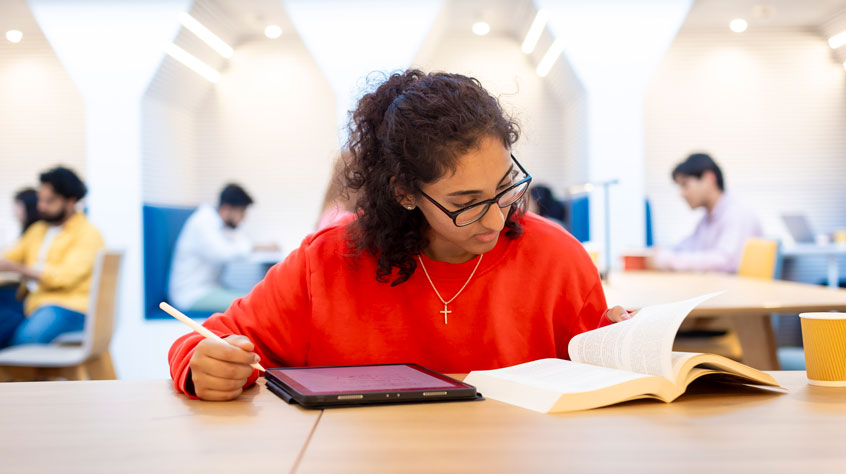 Student sitting in a study area, reading from a book and writing on an iPad.