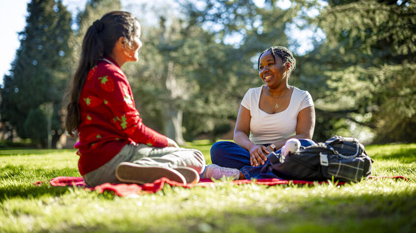 Two people sitting on a red blanket in a sunny park, smiling and talking.