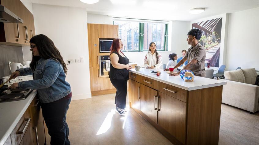 A group of six people socialize in a bright, modern kitchen. One person cooks while others chat at a central island.