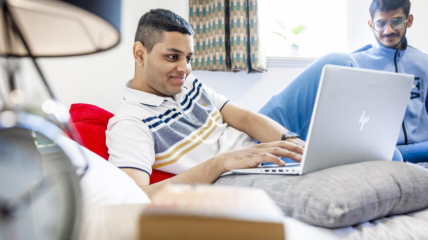 A young man in a striped polo shirt is smiling while using a laptop on a bed. Another person in blue is sitting nearby.