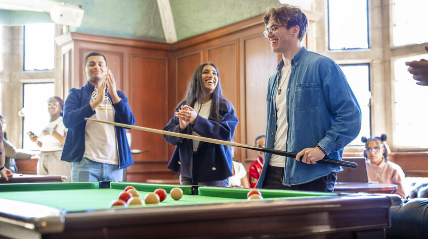 A group of young adults enjoy a lively moment around a billiard table in a sunlit room. One holds a cue stick, while others cheer and clap.