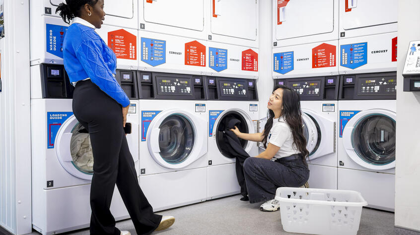 A woman in blue talks to another kneeling by a washing machine in a laundromat. Laundry basket in foreground.