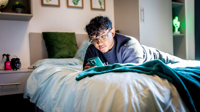 Man laying stomach down on a bed, looking at his phone.