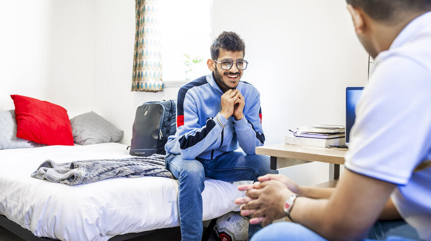Male students sitting on a bed and chair, talking, in student accommodation.