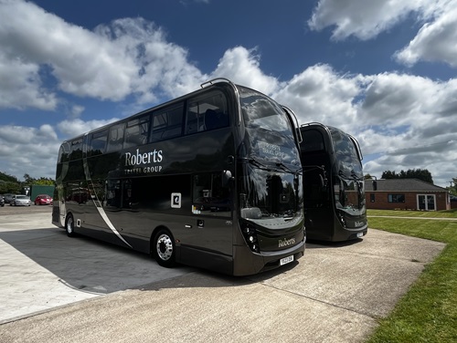 Two black double-decker buses parked side by side in a spacious parking lot under a clear blue sky.
