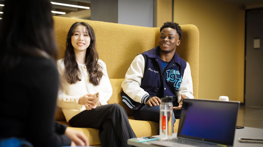 Three people sit on a yellow couch in a casual setting, smiling and engaged in conversation, with a laptop and water bottle on a table nearby.