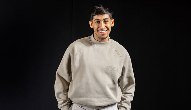 Person wearing a light grey sweatshirt, standing against a black studio background and facing the camera.