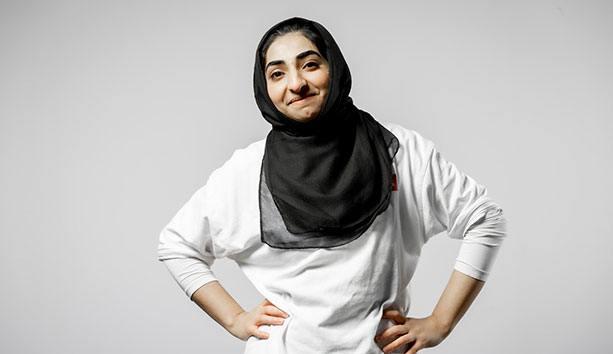 Person wearing a black headscarf and white top, standing with hands on hips against a light grey studio background.