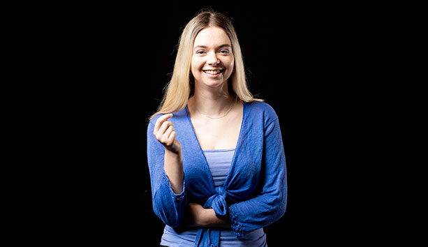 Person wearing a blue cardigan and top, standing against a black studio background and facing the camera.