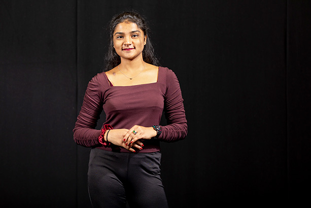 A young woman wearing a maroon top, smiling and standing against a neutral background.