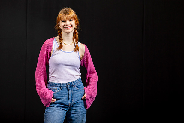 A young woman wearing a pink sweater stands smiling.