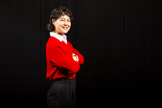 A young woman wearing a red sweater stands confidently, smiling at the camera.