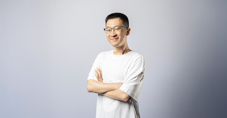 A young man wearing a white t-shirt and black pants, standing confidently with arms crossed.
