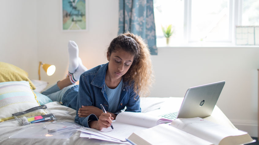 Student laying on a bed, writing in a notepad, with an open laptop next to her.