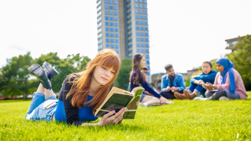 Female student sat outside on grass with a book