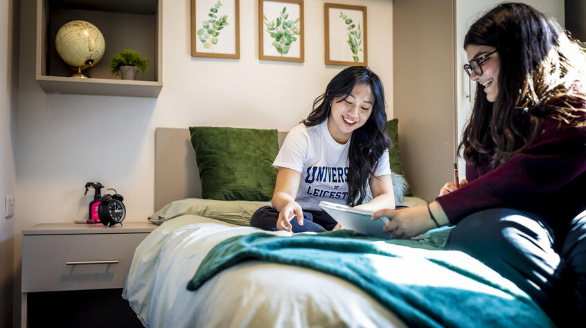 Two female students sitting on a bed in university accommodation room.