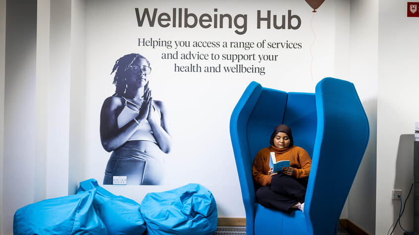 Female student sat on a comfy chair with a book in front of the wellbeing hub